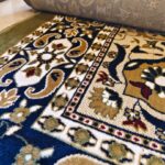 The Olive Dynasty Rug showcases a decorative design with floral and geometric patterns in blue, gold, and beige, paired with a partially rolled section.