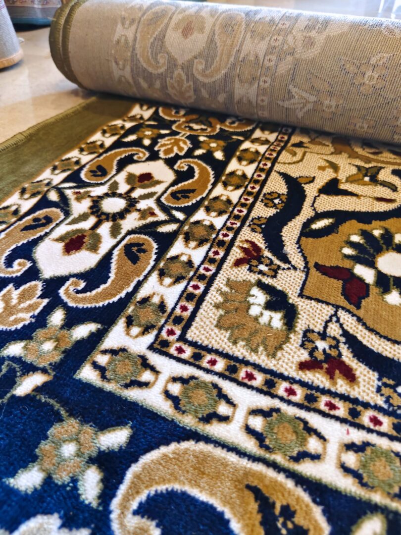 The Olive Dynasty Rug showcases a decorative design with floral and geometric patterns in blue, gold, and beige, paired with a partially rolled section.