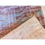 A close-up of the X Dime Abstract Rug, showing its multicolored design with red, brown, and light blue patterns, and a folded section revealing its beige underside.