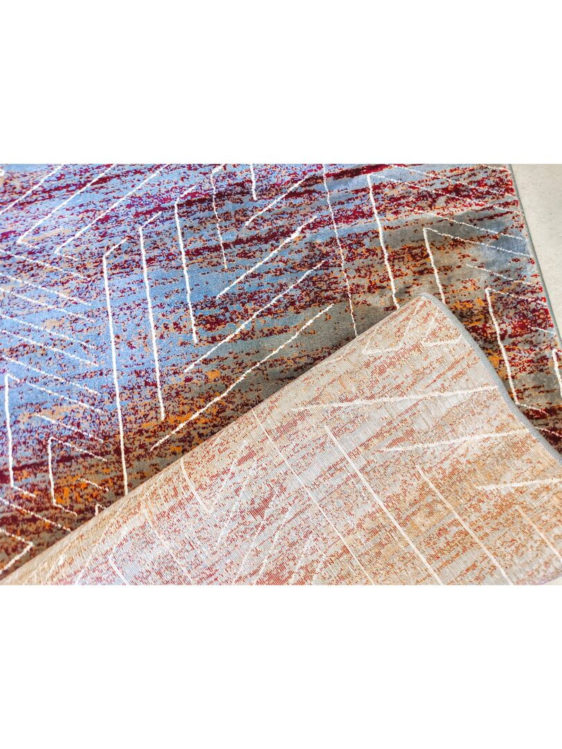 A close-up of the X Dime Abstract Rug, showing its multicolored design with red, brown, and light blue patterns, and a folded section revealing its beige underside.